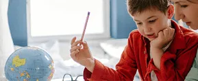 Niño concentrado escribiendo con un lápiz mientras una mujer lo acompaña y guía de cerca; en primer plano aparece un globo terráqueo sobre una mesa en un ambiente de estudio iluminado por luz natural.