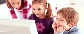 Tres niños pequeños observan con atención la pantalla de una computadora; dos están sentados frente al monitor mientras otro se asoma por detrás, todos en un entorno interior luminoso y enfocados en la actividad digital.