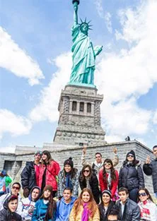 Grupo de jóvenes posando frente a la Estatua de la Libertad en Nueva York, con el monumento elevado sobre su base y el cielo parcialmente nublado de fondo durante una visita turística.